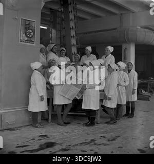 Slaviansk, Ukrainische SSR, UdSSR – 1981: Archivfoto einer großen Gruppe weiblicher Arbeiter in weißen Sanitärmänteln und Kopftüchern, die sich in der Fabrikhalle versammelten. Eine Frau liest ihren Kollegen während einer Pause die neuesten Nachrichten aus einer Zeitung vor. Diese Szene zeigt eine traditionelle „politische Informationsveranstaltung“, die ideologische Arbeit und den kollektiven Geist der Arbeiterklasse in der friedlichen Donbass-Region während der späten sowjetischen Ära beleuchtet. Stockfoto