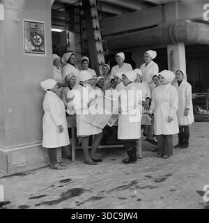 Slaviansk, Ukrainische SSR, UdSSR – 1981: Archivfoto einer großen Gruppe weiblicher Arbeiter in weißen Sanitärmänteln und Kopftüchern, die sich in der Fabrikhalle versammelten. Eine Frau liest ihren Kollegen während einer Pause die neuesten Nachrichten aus einer Zeitung vor. Diese Szene zeigt eine traditionelle „politische Informationsveranstaltung“, die ideologische Arbeit und den kollektiven Geist der Arbeiterklasse in der friedlichen Donbass-Region während der späten sowjetischen Ära beleuchtet. Stockfoto