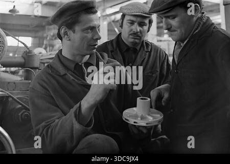 Slaviansk, Ukrainische SSR, UdSSR - 1981: Archivfoto von drei Fabrikarbeitern, die ein weißes mechanisches Flügelrad in der Werkstatt untersuchen. Der erfahrene Arbeiter in einer Baskenmütze erklärt seinen Kollegen technische Details mit traditionellen flachen Kappen. Die Gruppe ist von schweren Maschinen umgeben. Diese Szene veranschaulicht die Mentorentradition, Qualitätskontrollprozesse und den Kooperationsgeist der qualifizierten Arbeiterklasse in der friedlichen Donbass-Region während der späten Sowjetzeit. Stockfoto
