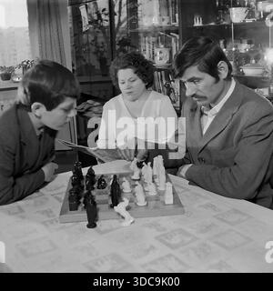 Slaviansk, Ukrainische SSR, UdSSR - 1981: Archiviertes Lifestyle-Foto, das einen sowjetischen Familienabend einfängt. Ein Vater mit Schnurrbart spielt Schach mit seinem Sohn an einem Tisch, während die Mutter im Hintergrund eine Zeitschrift liest. Das Zimmer verfügt über ein typisch spätsowjetisches Interieur, einschließlich einer polierten Wandeinheit („stenka“) mit Kristall und Büchern. Diese Szene spiegelt die Beliebtheit des Schachs und der inneren Stabilität in der friedlichen Donbass-Region während der späten sowjetischen Ära wider. Stockfoto