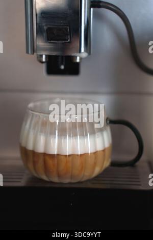 Glasbecher mit mehrschichtigem Kaffee mit Milchschaum unter silberner Espressomaschine in Kücheneinrichtung mit moderner und sanfter Beleuchtung, Ausstattung, Haushalt, schaumig, Stockfoto