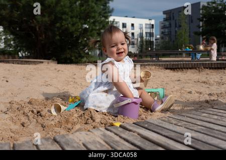 Ein lächelndes Kleinkind spielt in einem sonnigen Sandkasten mit Spielzeugen, umgeben von einem urbanen Park mit üppigem Grün, einem Holzweg und einem modernen Apartmentgebäude Stockfoto