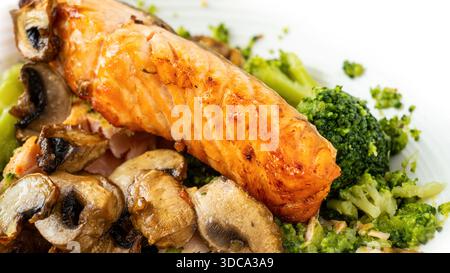 Ein Stück gegrillter Lachs wird auf einen Teller mit gekochten Pilzen und Brokkoli gelegt. Dieses Gericht wird während des Mittagessens an einem Esstisch serviert. Stockfoto
