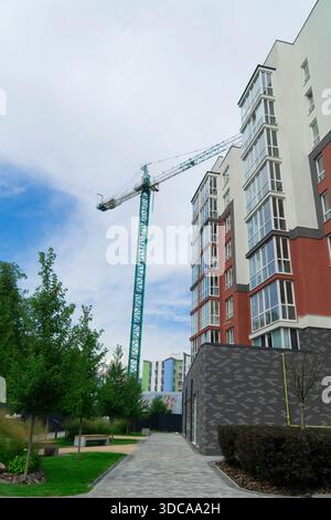 Neuer Wohnblock im Bau. Ein hoher grüner Turmkran steht neben einem modernen mehrstöckigen Gebäude mit einer Glasfassade vor einem bewölkten Himmel Stockfoto