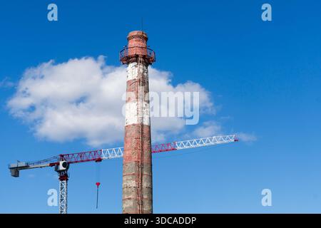 Hoher rot-weiß gestreifter Industrieschornstein neben einem modernen Turmkran unter klarem blauen Himmel mit kleinen weißen Wolken Stockfoto