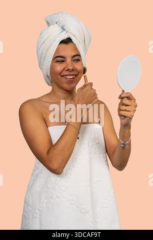 Frau im Handtuch, die eine Jadewalze auf ihr Gesicht aufträgt, während sie in einen Spiegel blickt Stockfoto