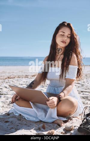 Frau in weißem Sommerkleid sitzt am Strand mit geschlossenem Laptop, entspannt, genießt die Sommersonne, abgelenkt von freiberuflicher Arbeit oder Studium Stockfoto