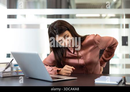 Junge Frau, die starke Rückenschmerzen und Unwohlsein empfindet, während sie an ihrem Schreibtisch sitzt und Probleme des sitzenden Lebensstils, der schlechten Körperhaltung und der gesundheitlichen Herausforderungen am Arbeitsplatz hervorhebt Stockfoto