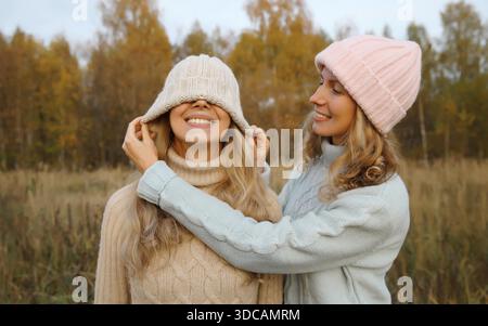 Porträt von zwei glücklichen Freundinnen, die Spaß haben, strickte Wintermütze über ihr Gesicht zu ziehen, die ihre Augen bedeckt, lustige Freundinnen, die draußen im Herbstpark zusammenkommen || Model veröffentlicht Stockfoto
