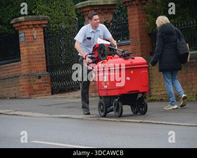 Ein Postbote von Royal Mail läuft eine Straße in Epping, Essex, UK, entlang und schiebt einen roten Postwagen Stockfoto