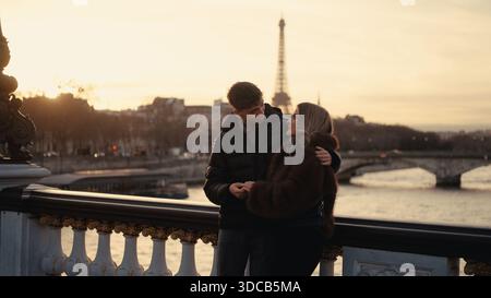Ein romantisches Paar umarmt sich auf einer Brücke in Paris Frankreich mit dem Eiffelturm, der bei Sonnenuntergang im Hintergrund sichtbar ist. Das Bild fängt einen Moment der Liebe ein Stockfoto