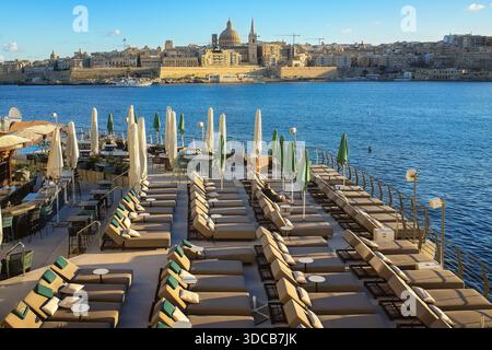 Luxuriöse, leere Strandterrasse bei Sonnenaufgang mit hübschen Reihen von Liegestühlen und geschlossenen Sonnenschirmen, mit malerischem Blick auf das Valletta. Stockfoto