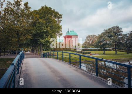 Eine breite Aufnahme einer Fußgängerbrücke mit blauem Geländer, die zu den mit Efeu bedeckten Backsteingebäuden und der grünen Kuppel des Krematoriums unter einem bedeckten Himmel führt. Stockfoto