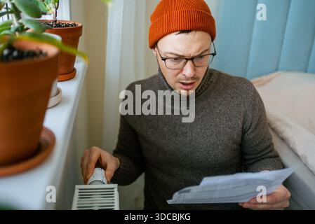 Mann mit orangefarbenem Hut und warmem Pullover, der einen Kühlerthermostat anpasst, schockiert und besorgt aussieht und eine riesige Stromrechnung hält, die steigende Lebenshaltungskosten während einer Energiekrise im Winter zeigt Stockfoto