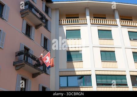 Eine helle Stadtfassade zeigt eine Schweizer Flagge, die von einem Balkon eines rosafarbenen Gebäudes mit grünen Fensterläden hängt, die europäische Architektur einfängt, in Farbtönen gehalten Stockfoto