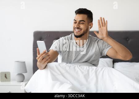 Fröhlicher Mann Aus Dem Nahen Osten, Der Videoanrufe Vom Bett Aus Macht, Während Er Die Virtuelle Verbindung Genießt Stockfoto