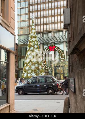 Geschmückter Weihnachtsbaum in einer Londoner Straßenszene vor einem Bürogebäude mit einem großen festlichen Kranz und einem schwarzen Taxi, das durch das Stadtzentrum führt. Stockfoto