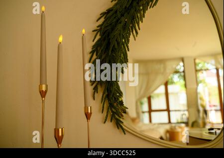 Hohe graue Kerzen in goldenen Haltern neben einem runden Spiegel, der mit Kiefernzweigen verziert ist. Konzept der minimalistischen Weihnachtsdekoration, festlich Stockfoto