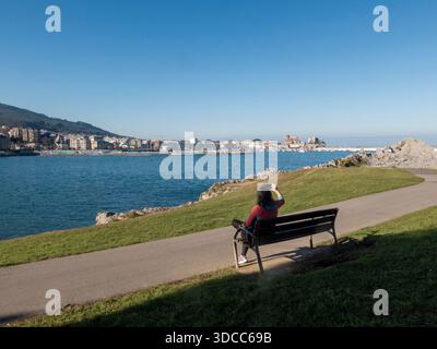 Frau, die auf einer Bank sitzt und auf die Stadt, den Hafen und das Meer in castro urdiales, spanien, blickt Stockfoto