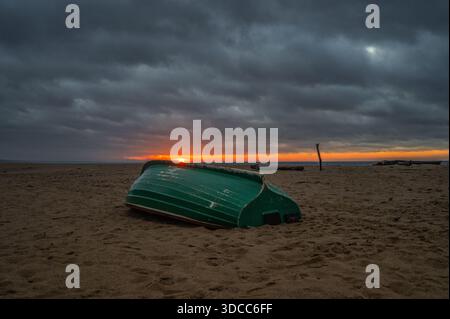 Stimmungsvolle Sonnenuntergangsszene mit türkisgrünem, umgedrehtem Holzboot am Sandstrand der Ostsee Stockfoto