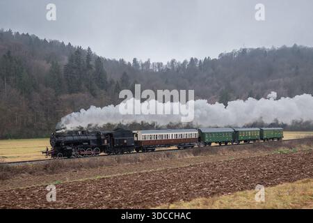 Eine klassische Dampflokomotive zieht klassische Personenwagen durch ein ruhiges ländliches Tal und gibt dicken weißen Dampf ab, wenn sie Felder und passiert Stockfoto