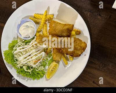 Goldbraune Fischfilets, die auf knusprigen, krinkle geschnittenen Pommes frites mit frischem Krautsalat und Zitronenkeil auf einem weißen Teller in gemütlicher Atmosphäre serviert werden. Stockfoto