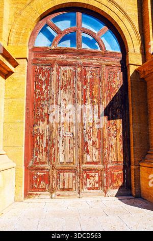 Verwitterte rote Holztür mit abblätternder Farbe steht innerhalb eines gelben Steinbogens, gekrönt von einem segmentierten Heckfenster. Alte Textur und rustikaler Charme Stockfoto