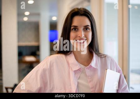 Lächelnde junge Frau mit Laptop in einem modernen Coworking-Raum, lässiges professionelles Porträt mit hellem Innenraum und natürlichem Licht Stockfoto
