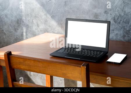 Laptop und Smartphone mit leeren weißen Bildschirmen auf einem Holzschreibtisch in einem modernen Arbeitsbereich. Betonwandhintergrund mit Sonnenlicht und Kopierraum, ideal für Stockfoto