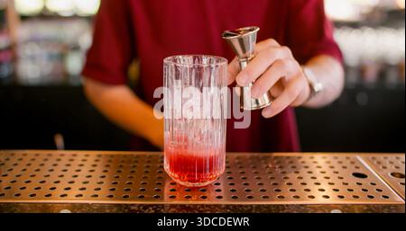 Cocktailzubereitung. Barservice. Barkeeper mit Jigger, der rote Flüssigkeit ins Glas gießt. Erfrischendes Getränk mit Eiswürfeln auf Metalltheke. Stockfoto