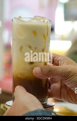Nahaufnahme einer Hand mit einem hohen Glas Eiskaffee mit cremigen Schichten, wirbelnden Texturen und sichtbaren Eiswürfeln in gemütlicher Atmosphäre. Stockfoto