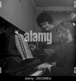 Slawjansk, Ukraine, UdSSR. 1976. Die stellvertretende Bashtovaya, Delegierte des 25. Kongresses der KPdSU, überwacht die Klavierpraxis ihrer Tochter zu Hause. Die Frau steht am Instrument und führt das Mädchen, das aus Notenblättern liest. Das polierte schwarze Klavier reflektiert das Gesicht der Mutter. Dieses Archivbild unterstreicht die Betonung der kulturellen Bildung und der inneren Harmonie der sowjetischen Arbeiterelite während der friedlichen Donbass-Ära. Die Tochter trägt eine große Schleife im Haar. Stockfoto