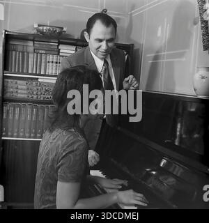 Slawjansk, Ukraine, UdSSR. 1976. Der Stellvertreter Rypalenko, Delegierter des 25. Kongresses der KPdSU, hört seiner Frau zuhause Klavier spielen zu. Der Mann im Anzug steht lächelnd am Instrument, während die Frau im gemusterten Kleid Musik spielt. Im Inneren befindet sich eine Bibliothek mit Büchern und einer Vase. Dieses Archivbild zeigt die kulturelle Freizeit, Familienharmonie und intellektuelle Atmosphäre der sowjetischen Arbeiterelite während der friedlichen Donbass-Ära. Stockfoto