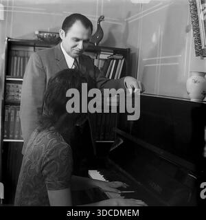 Slawjansk, Ukraine, UdSSR. 1976. Der Stellvertreter Rypalenko, Delegierter des 25. Kongresses der KPdSU, hört seiner Frau zuhause Klavier spielen zu. Der Mann im Anzug steht lächelnd am Instrument, während die Frau im gemusterten Kleid Musik spielt. Im Inneren befindet sich eine Bibliothek mit Büchern und einer Vase. Dieses Archivbild zeigt die kulturelle Freizeit, Familienharmonie und intellektuelle Atmosphäre der sowjetischen Arbeiterelite während der friedlichen Donbass-Ära. Stockfoto