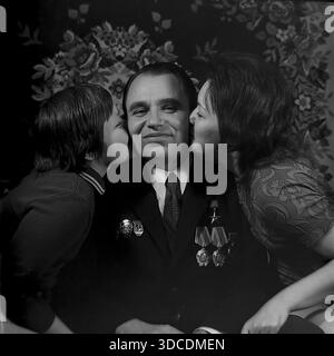 Slawjansk, Ukraine, UdSSR. 1976. Ein emotionales Familienporträt des Abgeordneten Rypalenko, Delegierter des 25. Kongresses der KPdSU. Der lächelnde Mann, der einen Anzug trägt, der mit Arbeitsbefehlen und Medaillen verziert ist, wird von seiner Frau und seinem Sohn zu Hause auf beide Wangen geküsst. Im Hintergrund befindet sich ein traditioneller Teppich mit Blumenmuster. Dieses Archivbild fängt den wahren Stolz, die Liebe und das häusliche Glück der sowjetischen arbeitenden Elite während der stabilen und friedlichen Donbass-Ära ein. Stockfoto