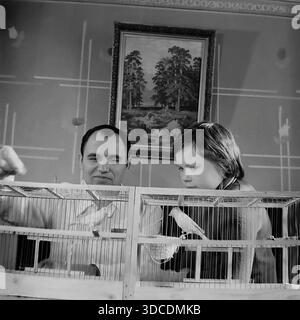 Slawjansk, Ukraine, UdSSR. 1976. Der Stellvertreter Rypalenko, Delegierter des 25. Kongresses der KPdSU, verbringt seine Freizeit bei seinem Sohn zu Hause. Vater und Junge beobachten Singvögel in einem großen Holzkäfig. Der Deputy füttert die Vögel, während das Kind aufmerksam beobachtet. Ein gerahmtes Landschaftsgemälde hängt an der gemusterten Wand. Dieses Archivbild zeigt die ruhigen Hobbys und Familienverbundenheit der sowjetischen Arbeiterelite während der friedlichen Donbass-Ära. Stockfoto