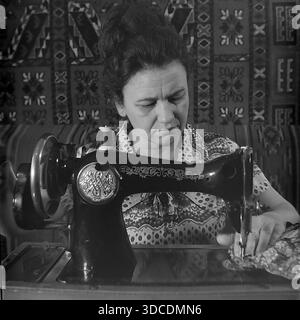 Slawjansk, Ukraine, UdSSR. 1976. Der Stellvertreter Kalaschnikowa, Delegierte des 25. Kongresses der KPdSU, beschäftigt sich mit der Handarbeit zu Hause. Die Frau sitzt an einer Vintage-schwarzen Handnähmaschine mit goldenen Blumenornamenten, die sorgfältig Stoff nähen. Ein traditioneller Teppich hängt im Hintergrund an der Wand. Dieses Archivbild veranschaulicht das häusliche Leben, den Fleiß und die traditionellen Haushaltsfähigkeiten der sowjetischen Arbeiterelite während der stabilen und friedlichen Donbass-Ära. Stockfoto