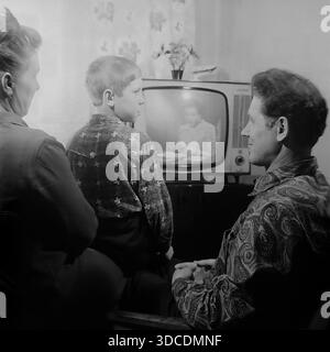 Slawjansk, Ukraine, UdSSR. 1976. Der Stellvertreter Schestopalov, Delegierter des 25. Kongresses der KPdSU, schaut mit seiner Familie zu Hause fern. Der Mann, der ein modisches Paisley-Hemd trägt, seine Frau und ihr Sohn versammeln sich um ein Vintage-Röhrenfernsehgerät mit einer Zimmerpflanze oben. Der Junge sieht seinen Vater an, während er vor dem Bildschirm sitzt. Dieses Archivbild fängt die typische abendliche Freizeit und häusliche Atmosphäre der sowjetischen Arbeiterelite während der friedlichen Donbass-Ära ein. Stockfoto