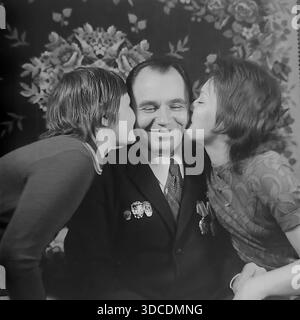 Slawjansk, Ukraine, UdSSR. 1976. Ein emotionales Familienporträt des Abgeordneten Rypalenko, Delegierter des 25. Kongresses der KPdSU. Der lächelnde Mann, der einen Anzug trägt, der mit Arbeitsbefehlen und Medaillen verziert ist, wird von seiner Frau und seinem Sohn zu Hause auf beide Wangen geküsst. Im Hintergrund befindet sich ein traditioneller Teppich mit Blumenmuster. Dieses Archivbild fängt den wahren Stolz, die Liebe und das häusliche Glück der sowjetischen arbeitenden Elite während der stabilen und friedlichen Donbass-Ära ein. Stockfoto