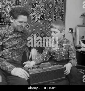 Slawjansk, Ukraine, UdSSR. 1976. Der Stellvertreter Schestopalov, Delegierter des 25. Kongresses der KPdSU, verbringt Freizeit mit seinem Sohn zu Hause. Der Vater und der Junge passen die Bedienelemente eines tragbaren Recorders auf Rollen vor dem Hintergrund eines gemusterten Wandteppichs an. Der Mann trägt ein modisches Paisley-Hemd. Dieses Archivbild fängt das Interesse an Technologie und warmen Familienmomenten der sowjetischen Arbeiterelite während der stabilen und friedlichen Donbass-Ära ein. Stockfoto