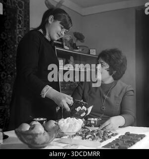 Slawjansk, Ukraine, UdSSR. 1976. Die stellvertretende Bashtovaya, Delegierte des 25. Kongresses der KPdSU, genießt zu Hause eine Teeparty mit ihrer Tochter. Das Mädchen gießt Tee aus einem Keramiktopf. Der Tisch ist voller Leckereien: Eine Schüssel mit frischen Äpfeln, Zuckerklumpen und Schokolade. Ein Sideboard mit Figuren und ein Wandteppich runden das gemütliche Interieur ab. Dieses Archivbild veranschaulicht den häuslichen Komfort und Wohlstand der sowjetischen Arbeiterelite während der stabilen, friedlichen Donbass-Ära. Stockfoto