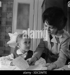 SLAWJANSK, UKRAINE, UdSSR – 1976: Die stellvertretende Ladygina, eine angesehene Arbeiterin des Slawjansk Soda Combine und Delegierte des XXV KPdSU-Kongresses, hilft ihrer Tochter bei den Hausaufgaben. Die aufmerksame Mutter zeigt auf Texte in einem Buch, während das Mädchen mit einer großen, ikonischen weißen Schleife vorsichtig auf den Heimtisch zusieht. Ein Archivbild, das frühe Bildung, Alphabetisierung und engagierte mütterliche Betreuung in der friedlichen Donbass-Region während der stabilen sowjetischen Breschnew-Ära veranschaulicht. Stockfoto