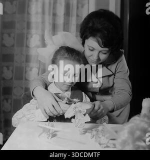SLAWJANSK, UKRAINE, UdSSR - 1976: Die stellvertretende Ladygina, eine angesehene Arbeiterin des Slawjansk Soda Combine und Delegierte des XXV KPdSU-Kongresses, genießt kreative Freizeit mit ihrer Tochter. Die lächelnde Mutter sieht zu, wie das Mädchen mit einer großen ikonischen Schleife fleißig Papierfiguren mit einer Schere am Tisch ausschneidet. Ein Archivbild, das Familienhobbys, die Entwicklung von Kindern und das häusliche Glück in der friedlichen Donbass-Region während der stabilen sowjetischen Breschnew-Ära veranschaulicht. Stockfoto