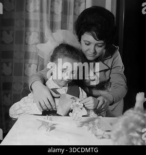 SLAWJANSK, UKRAINE, UdSSR - 1976: Die stellvertretende Ladygina, eine angesehene Arbeiterin des Slawjansk Soda Combine und Delegierte des XXV KPdSU-Kongresses, genießt kreative Freizeit mit ihrer Tochter. Die lächelnde Mutter sieht zu, wie das Mädchen mit einer großen ikonischen Schleife fleißig Papierfiguren mit einer Schere am Tisch ausschneidet. Ein Archivbild, das Familienhobbys, die Entwicklung von Kindern und das häusliche Glück in der friedlichen Donbass-Region während der stabilen sowjetischen Breschnew-Ära veranschaulicht. Stockfoto