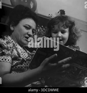 Slawjansk, Ukraine, UdSSR. 1976. Die Abgeordnete Kalaschnikowa, Delegierte des 25. Kongresses der KPdSU, liest ihrer Tochter das ukrainische Buch "Siebenhundert Nachtigallen" vor. Die Mutter und das Mädchen sind in Nahaufnahme vor dem Hintergrund eines Sideboards mit Kristallgläsern abgebildet. Die Frau erklärt die Geschichte. Dieses Archivbild fängt die Betonung auf Bildung, kulturelles Erbe und häuslichem Komfort der sowjetischen Arbeiterelite während der stabilen und friedlichen Donbass-Ära ein. Stockfoto