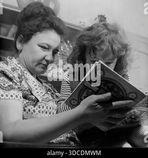 Slawjansk, Ukraine, UdSSR. 1976. Die Abgeordnete Kalaschnikowa, Delegierte des 25. Kongresses der KPdSU, liest ihrer Tochter das ukrainische Buch "Siebenhundert Nachtigallen" vor. Die Mutter und das Mädchen sind in Nahaufnahme vor dem Hintergrund eines Sideboards mit Kristallgläsern abgebildet. Die Frau erklärt die Geschichte. Dieses Archivbild fängt die Betonung auf Bildung, kulturelles Erbe und häuslichem Komfort der sowjetischen Arbeiterelite während der stabilen und friedlichen Donbass-Ära ein. Stockfoto
