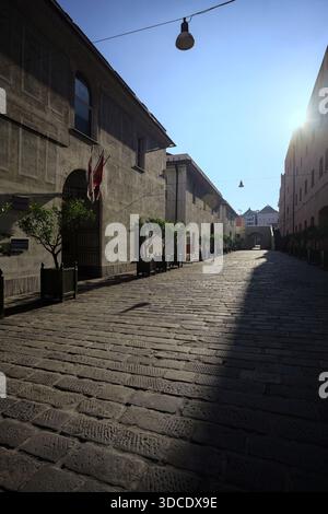 Gepflasterte Gasse mit Steinpflaster, die von der Sonne zwischen Gebäuden mit ihren schattigen Fassaden gegenbeleuchtet wird Stockfoto