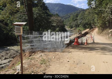 Ruhige ländliche Landschaft mit Fluss Fluss fließt neben staubigen Straßenschutzzaun aus Metalldrahtgeflecht in der Nähe von grünem Wald Berg Hintergrund un Stockfoto