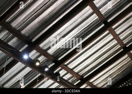 Flacher Blick auf das moderne Wellblechdach mit brauner Holzbalkenstruktur und heller Glühbirne hängender dunkler industrieller Deckenkonstruktion Stockfoto