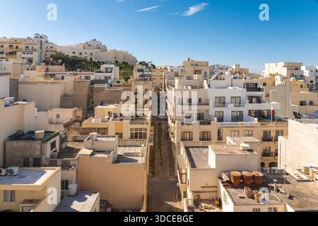Blick auf sonnendurchflutete Gebäude, die lange Schatten in einer engen Straße unter einem hellblauen Himmel werfen und eine warme, einladende Szene schaffen, Mellieha, Malta. Stockfoto